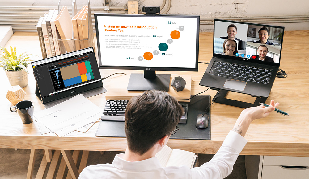 A professional working at a desk with multiple devices, including a desktop monitor displaying a presentation, a laptop showing a video conference, and a portable screen with analytics data.