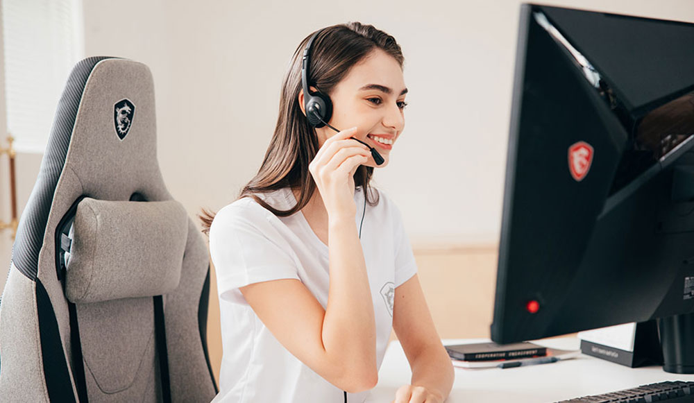 A woman wearing a headset and smiling while speaking into a microphone, sitting at a desk with a computer monitor and keyboard.